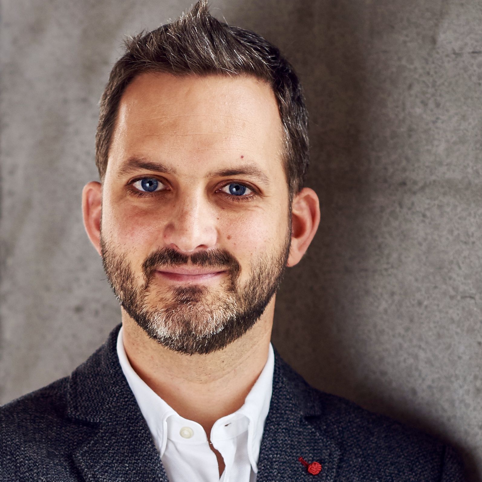 Geschäftsporträt eines Mannes in einem grauen Sakko mit weißem Hemd vor einer Betonwand.