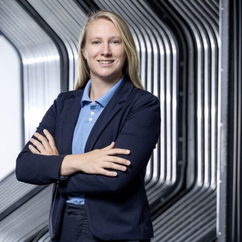Blonde business professional in a navy blazer standing with folded arms in a modern industrial corridor.
